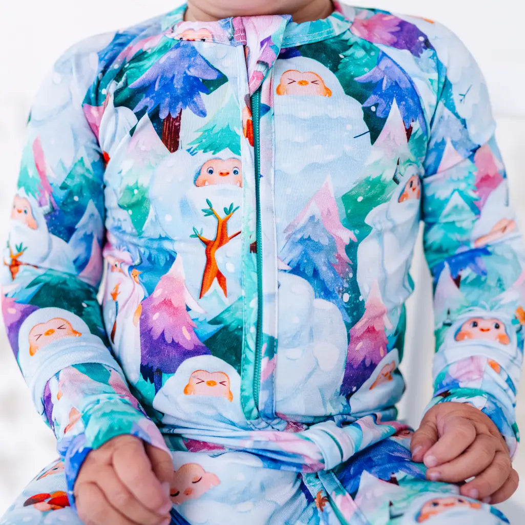 Close up of a toddler sitting with hands in the lap in a zip up pajama with a print of friendly white yetis among teal, blue, and purple pine trees, the front zipper running down the center.